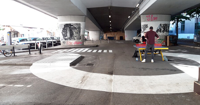 Blick auf eine Zwischennutzungsfläche unter einer Straßenbrücke mit bemalten Brückenpfeilern, Tischtennisplatte und markierten Fußgängerwegen.