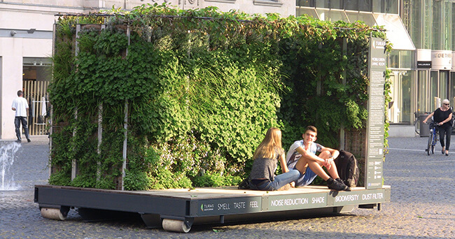 Mobiles grünes Zimmer mit bepflanzten Wänden und integrierter Sitzfläche auf einem Platz in der Stadt. Zwei Personen sitzen auf der Bank vor der begrünten Konstruktion.