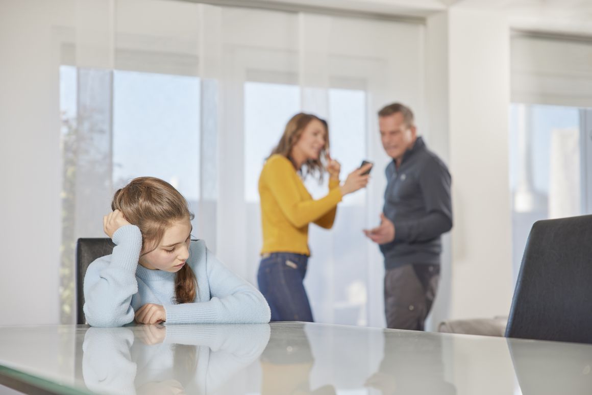 Eltern diskutieren laut und die Tochter sitzt währenddessen traurig am Tisch.